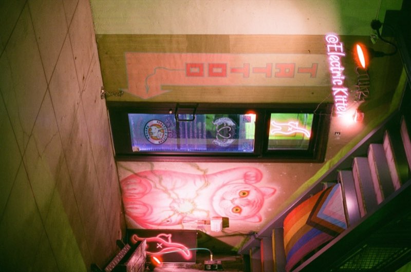 A tattoo shop stairwell: neon cat sign, painted mural, pride flag on the rail.